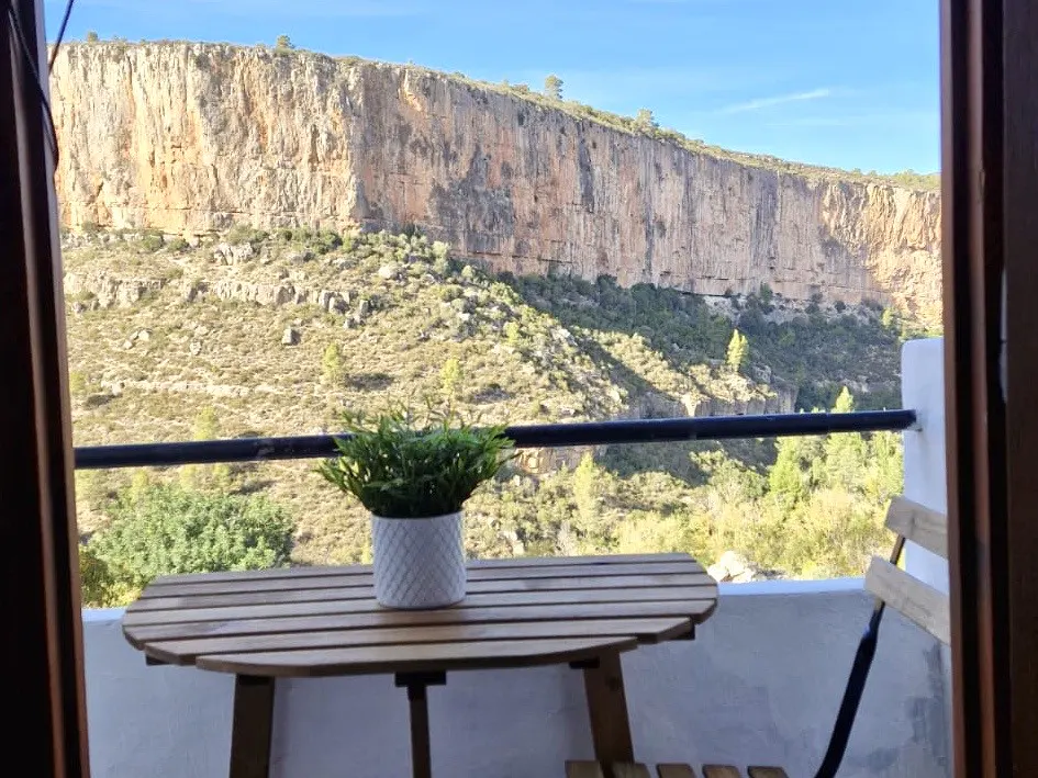 View from a balcony with a wooden table, chair, and potted plant, overlooking a valley and a large cliff face.