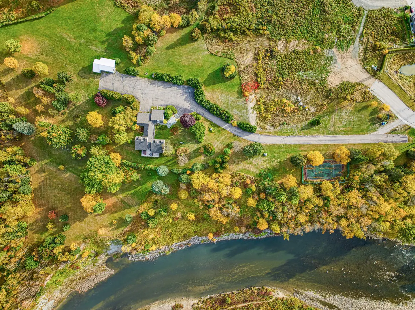 Aerial view of a house with a tennis court next to a river surrounded by colorful trees.
