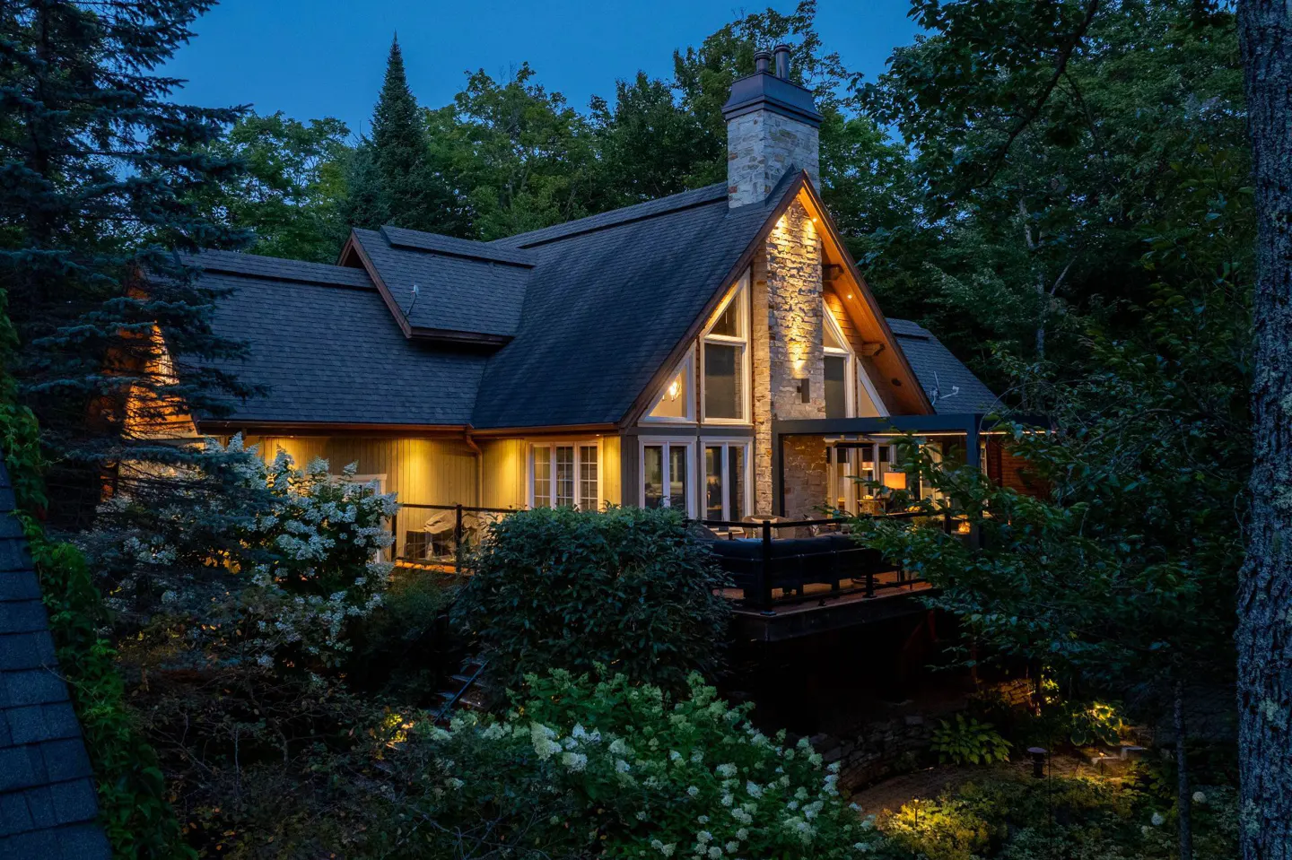 A-frame house with stone chimney, dark roof, and deck, nestled among trees at dusk. Lights glow from windows and landscape.