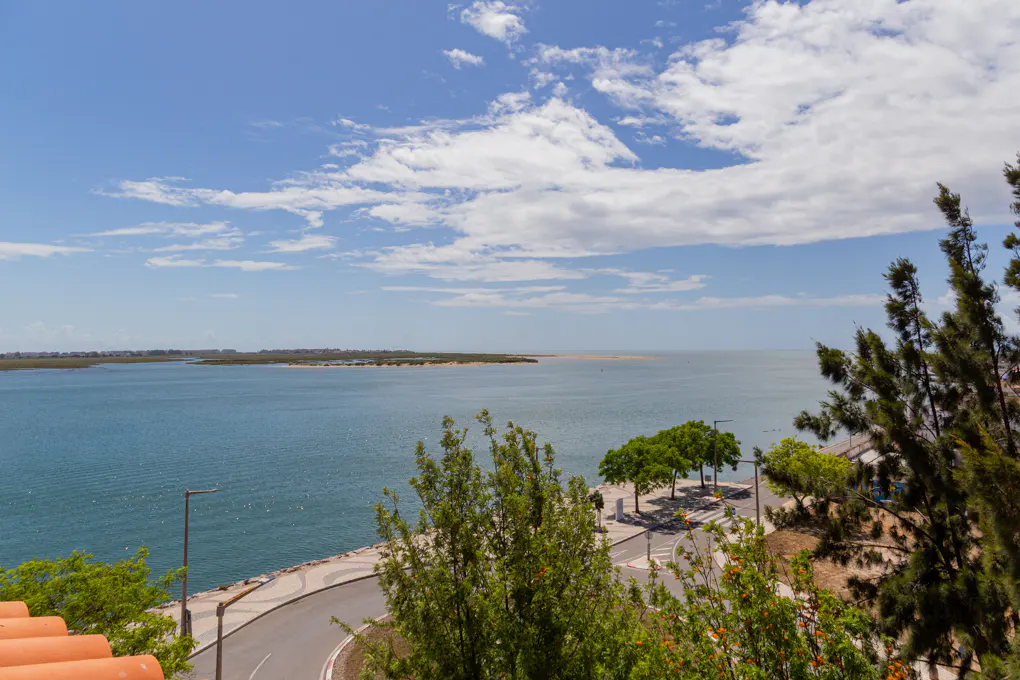Scenic view of a blue bay under a partly cloudy sky, with a road, trees, and a glimpse of orange roof tiles in the foreground.