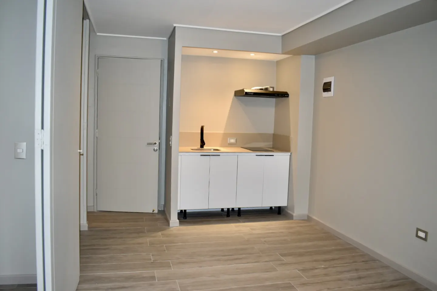 A studio apartment with gray walls, light wood floors, and a small white kitchen with black accents.