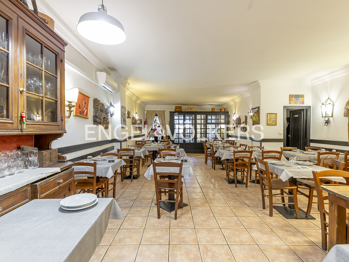 Restaurant interior with wooden tables and chairs, set with plates and glasses. Tiled floor and white walls with art.