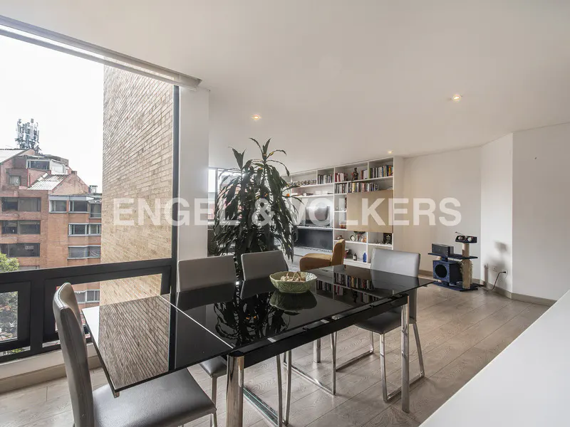 Bright dining area with a black table, gray chairs, and a large window overlooking a city. A bookshelf and a plant are in the background.