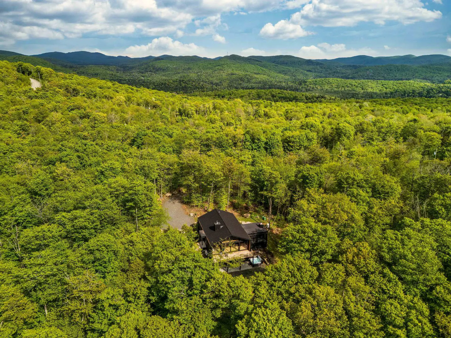 Aerial view of a modern black cabin nestled in a lush green forest with rolling mountains under a blue sky.
