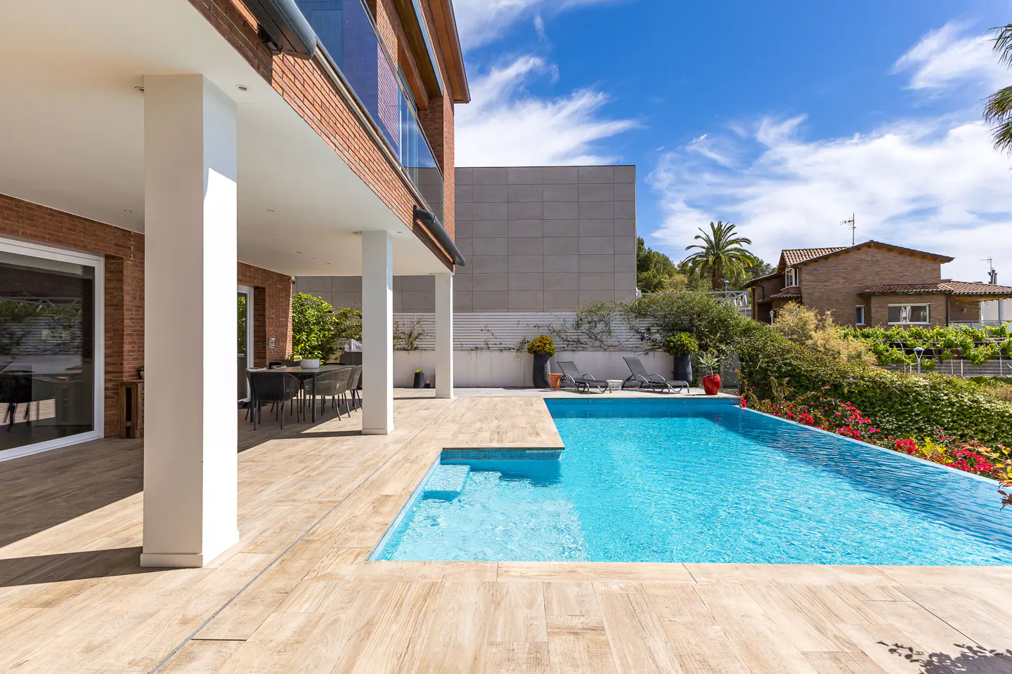 Outdoor view of a modern home with a blue swimming pool, patio furniture, and a brick exterior under a partly cloudy sky.