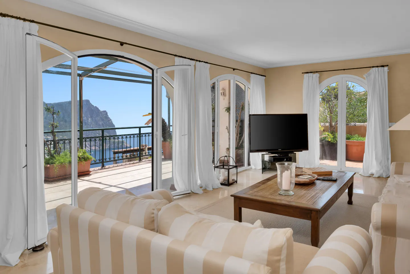 Living room with striped sofa, wooden coffee table, and open arched doors leading to a balcony with a scenic ocean view.