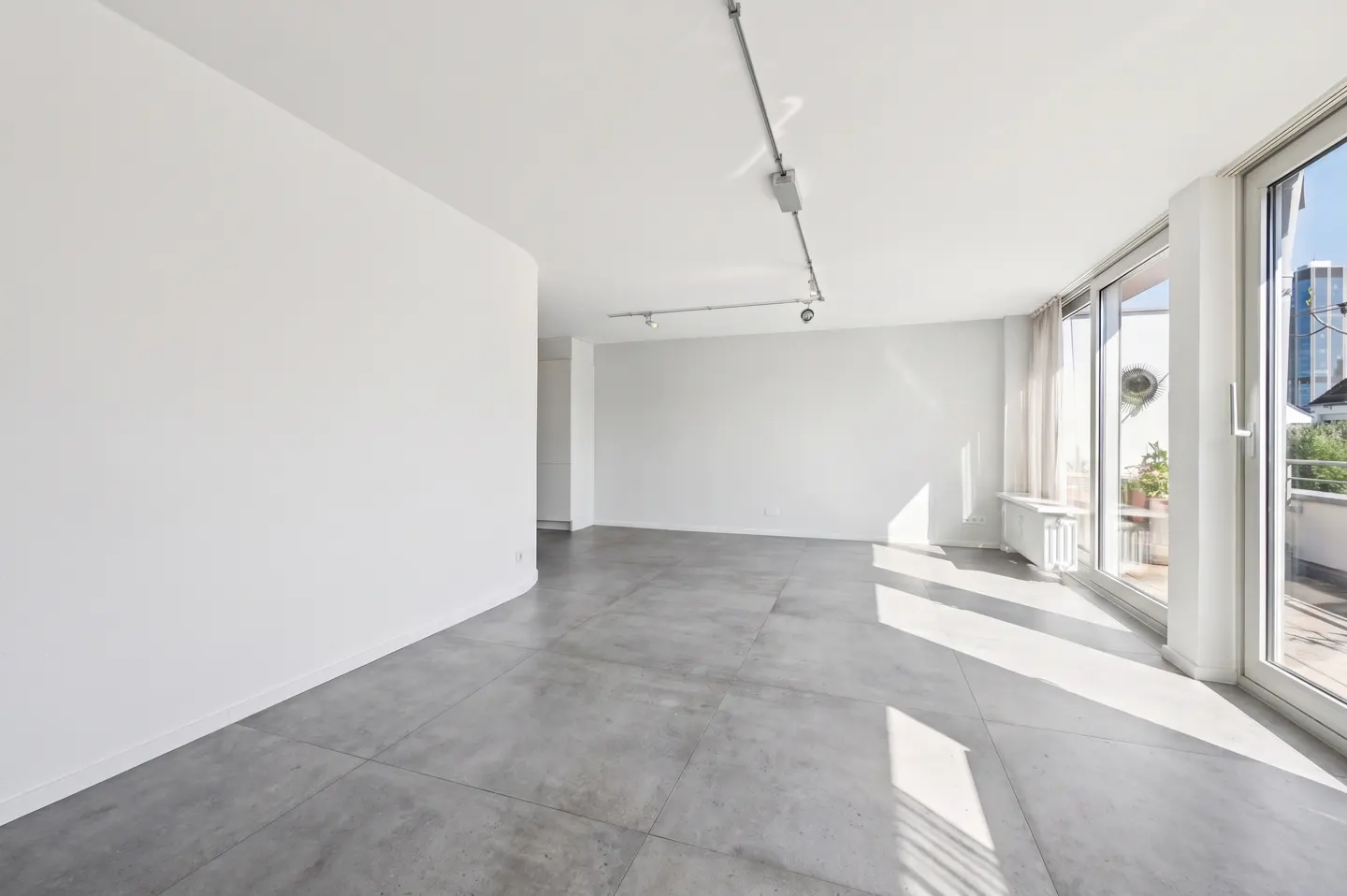 Bright, empty room with white walls, gray tile floor, and sliding glass doors to a balcony. Track lighting on the ceiling.