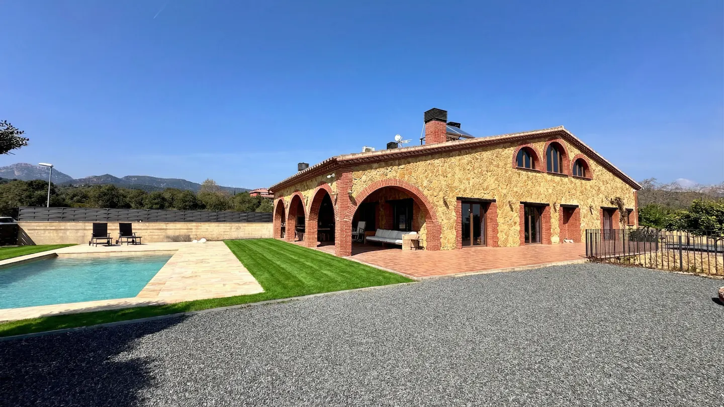Exterior view of a stone house with red brick arches, a pool, and a gravel driveway on a sunny day.