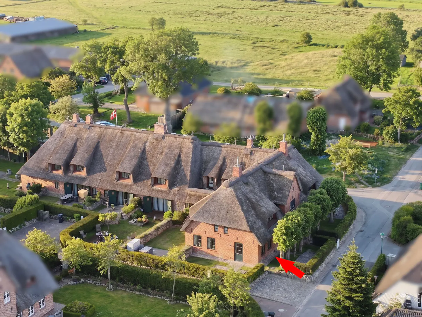 Aerial view of a brick house with a thatched roof, surrounded by green lawns and trees. An arrow points to the entrance.
