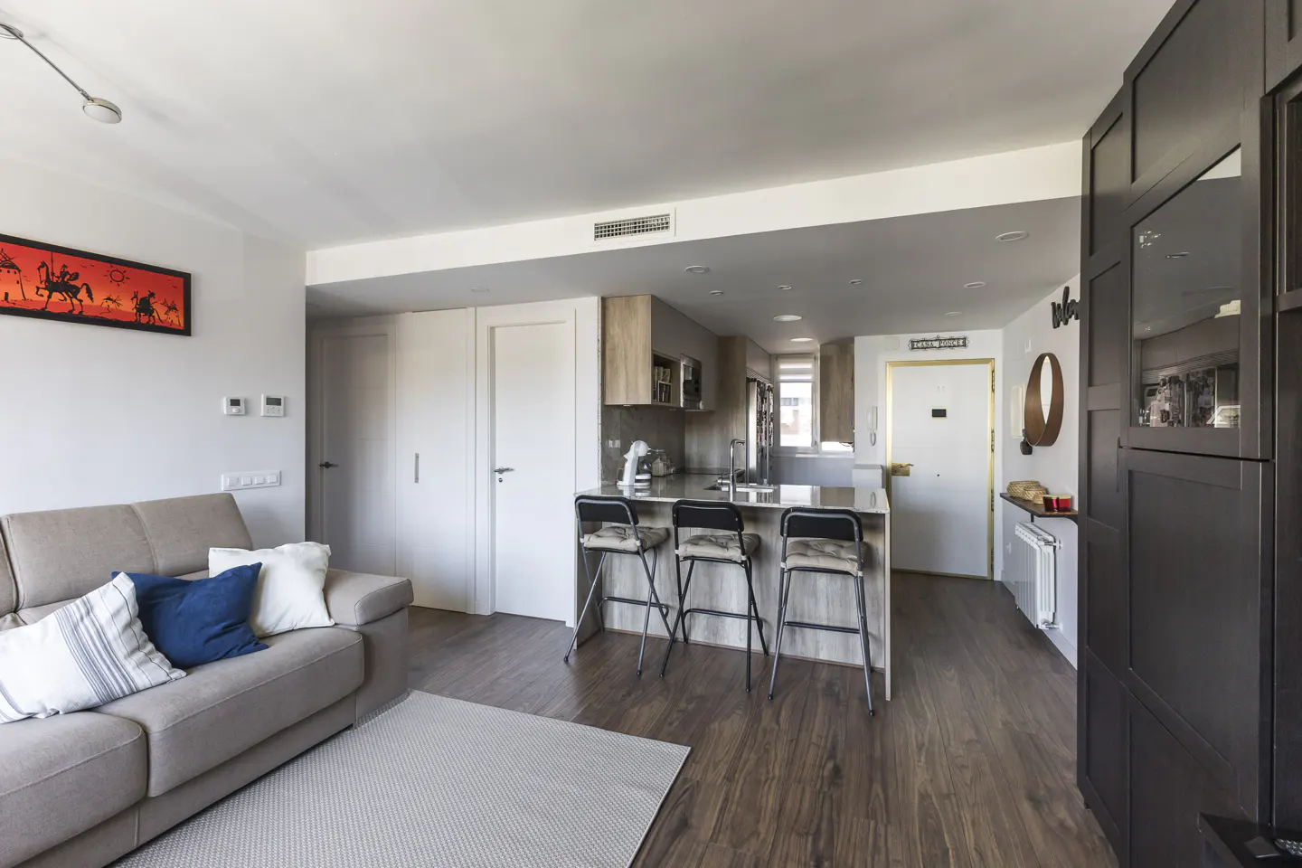 A bright, modern apartment living space with a gray sofa, kitchen island with stools, and dark wood floors.