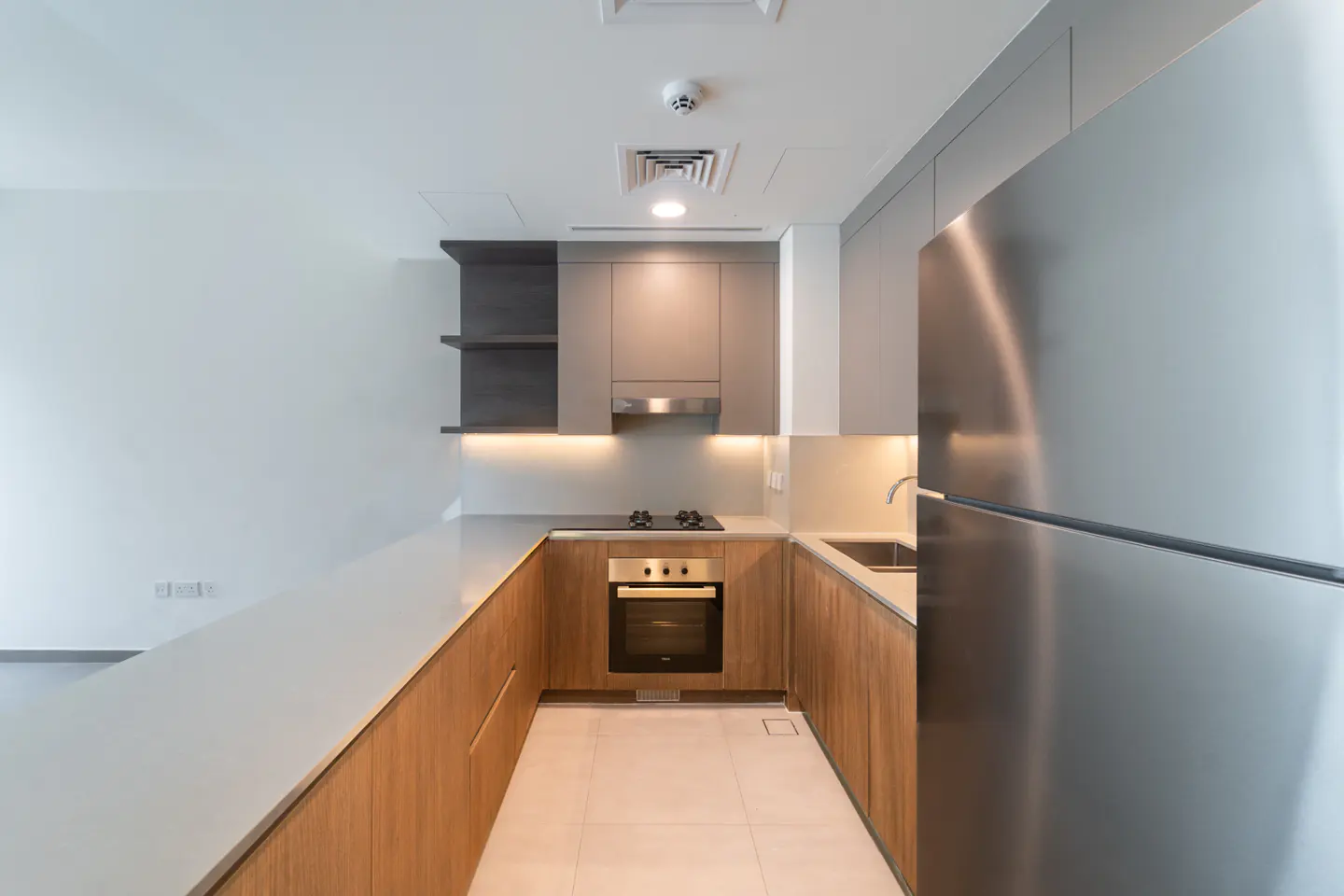 A modern kitchen with wood cabinets, stainless steel appliances, and a white countertop. There is an oven, cooktop, and sink.