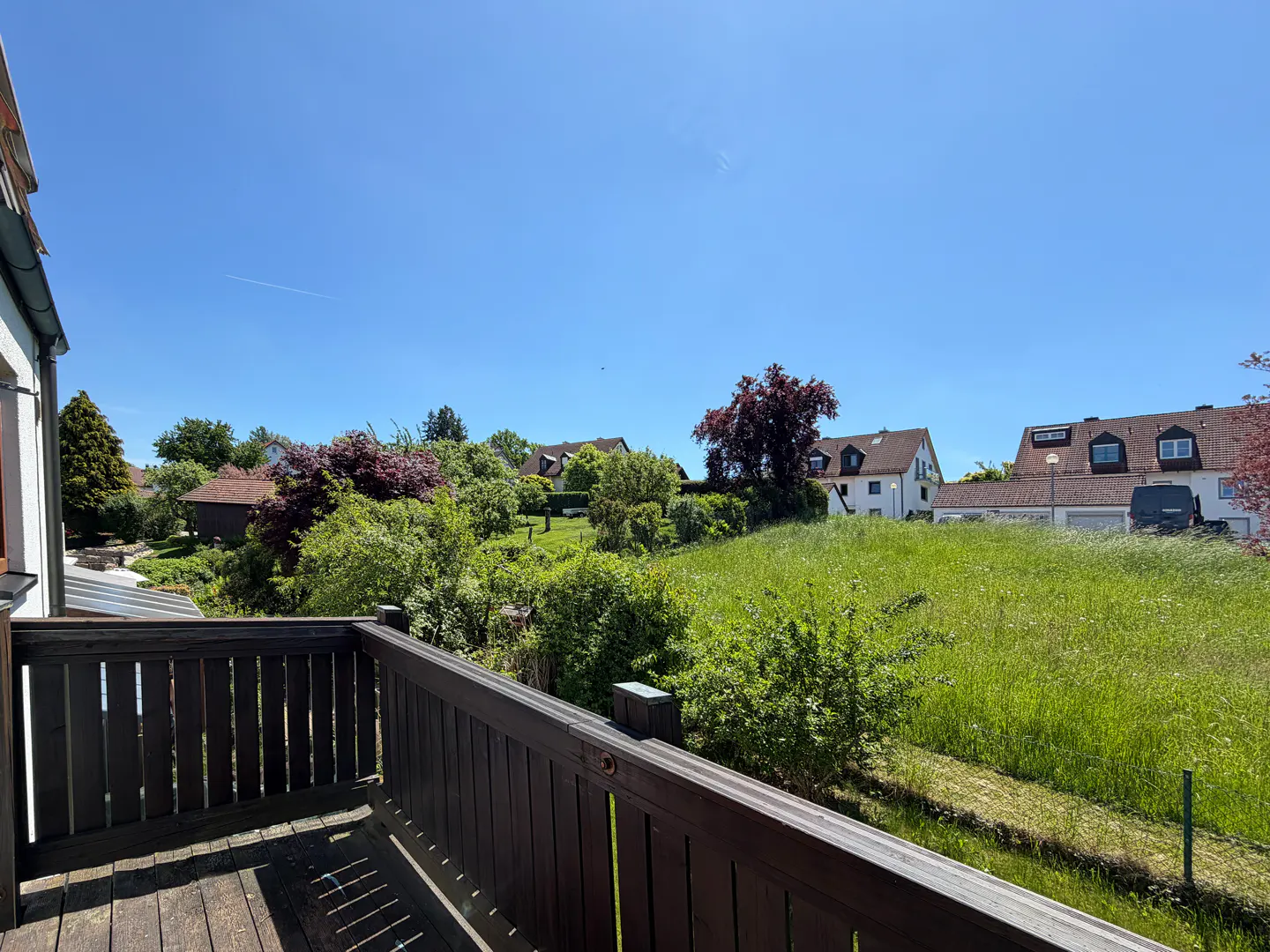 View from a wooden balcony overlooking a lush green meadow, trees, and residential houses under a clear blue sky.