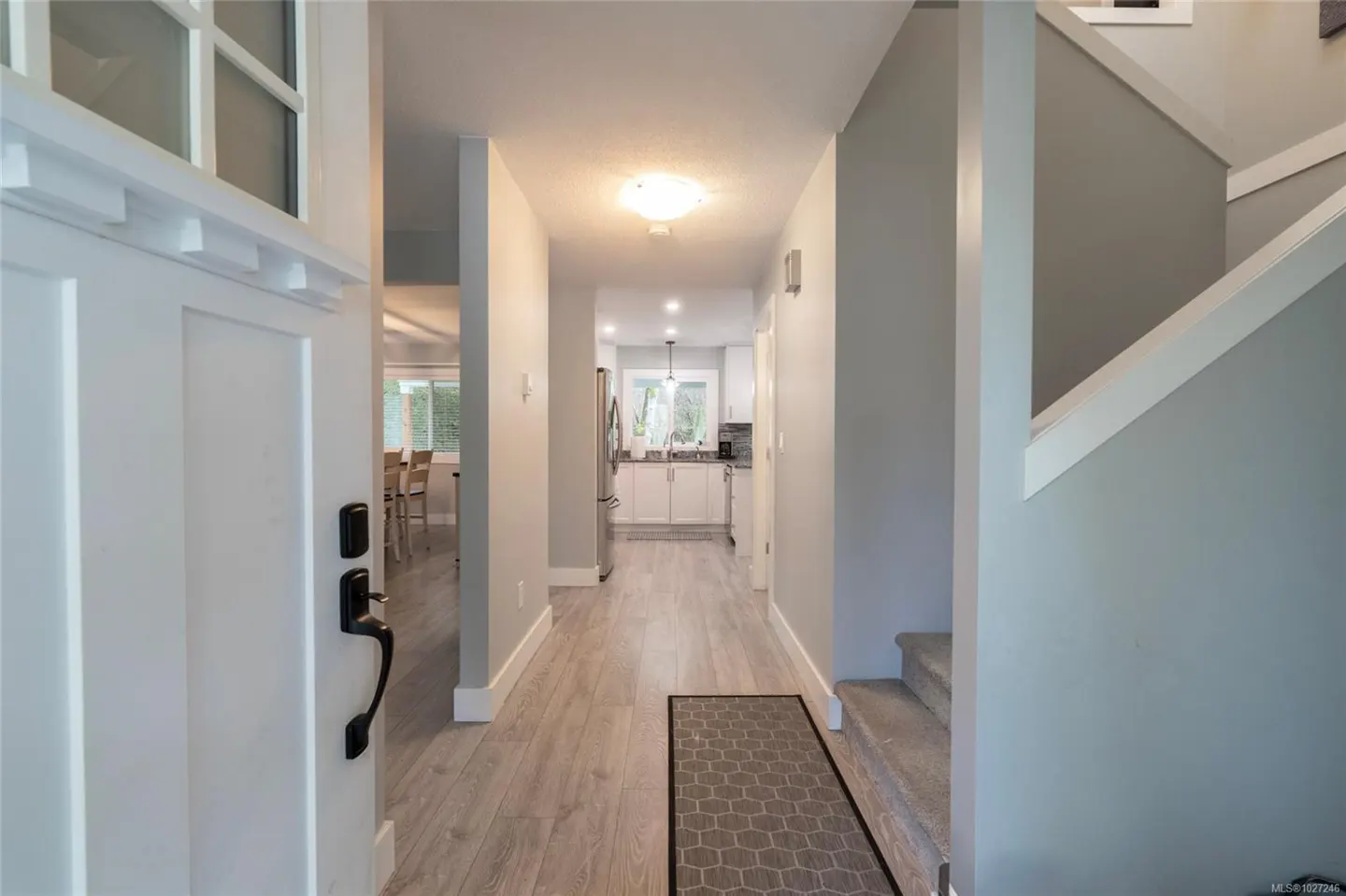 Open white front door reveals a hallway with light gray walls and wood floors leading to a kitchen. Stairs are on the right.