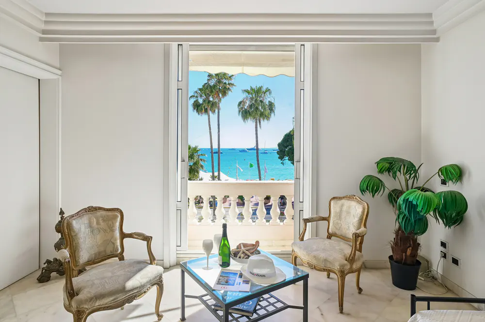 Bright living room with two ornate chairs, a glass table with wine, and a balcony overlooking a blue ocean with palm trees.