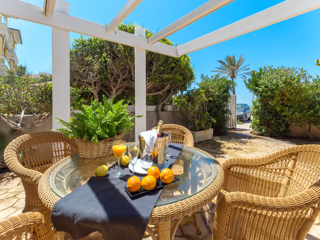 Outdoor patio with wicker furniture, glass table, and refreshments under a white pergola. Lush greenery and blue sky in the background.