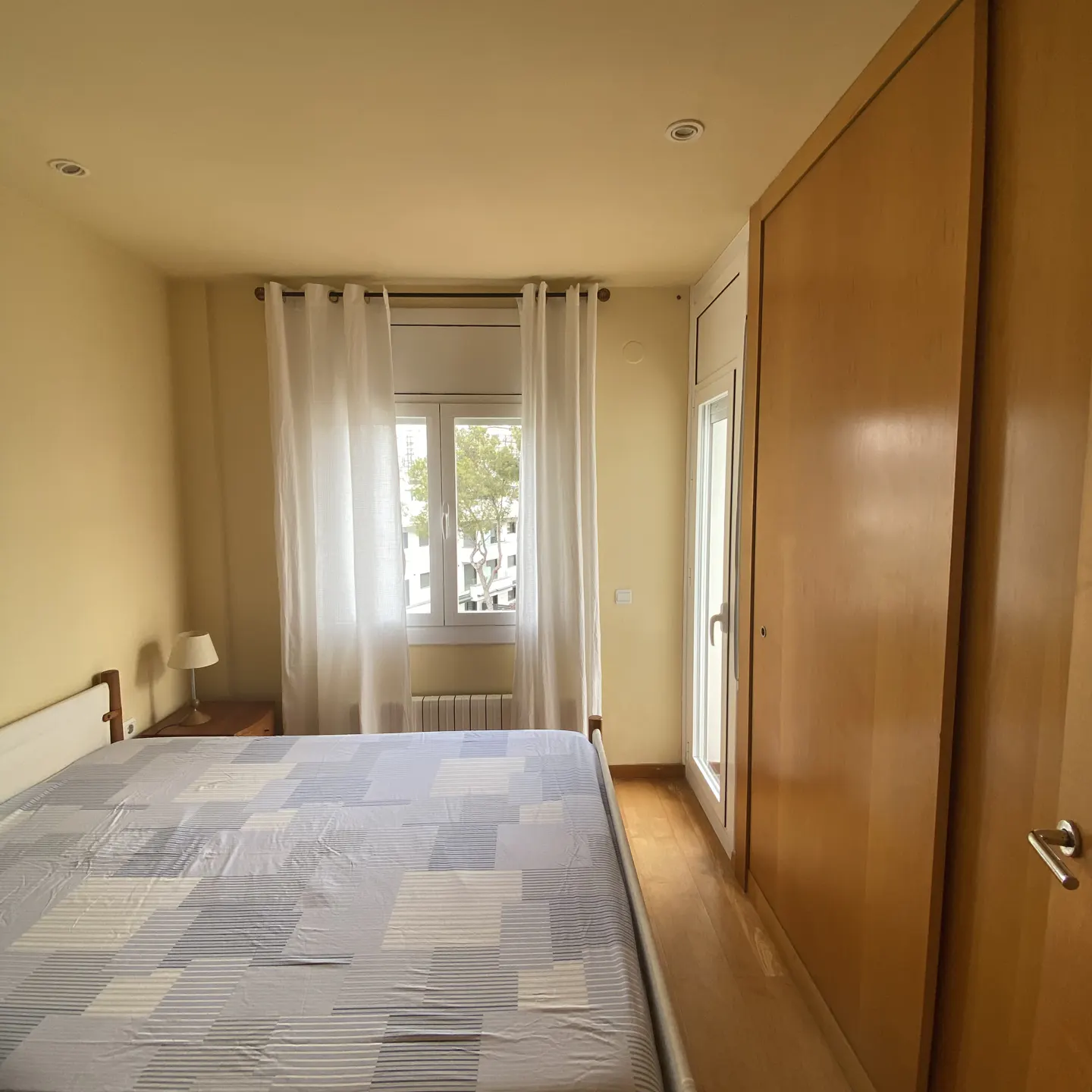 Bedroom with a bed covered in a blue and white patterned sheet, a window with white curtains, and a wooden wardrobe.