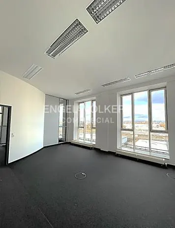 Bright, empty office space with black carpet, white walls, and large windows overlooking a cityscape. Overhead fluorescent lighting.