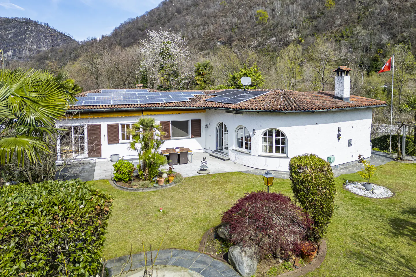 Aerial view of a white house with solar panels, a red tile roof, and a green lawn with trees and shrubs. A Swiss flag flies in the background.