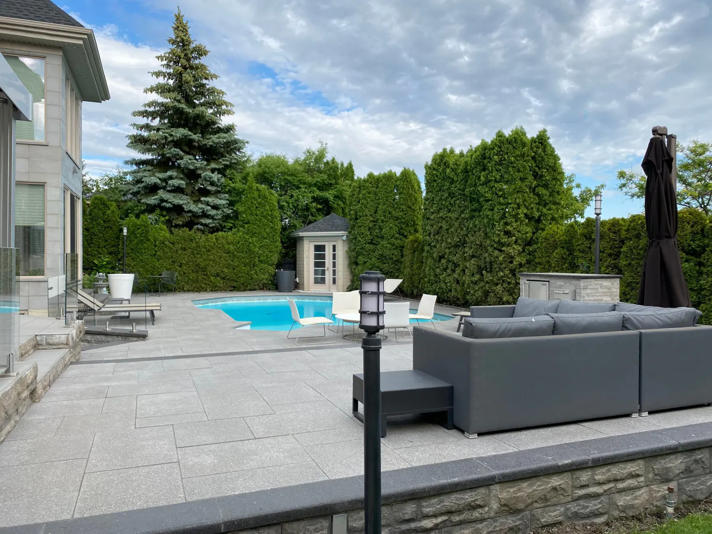 Backyard with a blue pool, gray patio, white chairs, and a gray sofa. Green trees and a small shed are in the background.