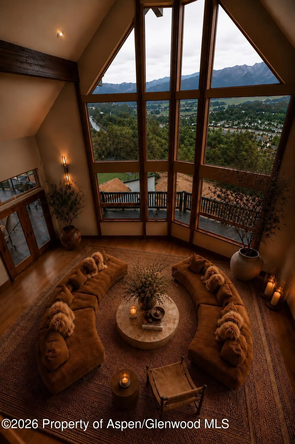 A living room with a view features curved brown sofas, a round table, and floor-to-ceiling windows overlooking a green valley.