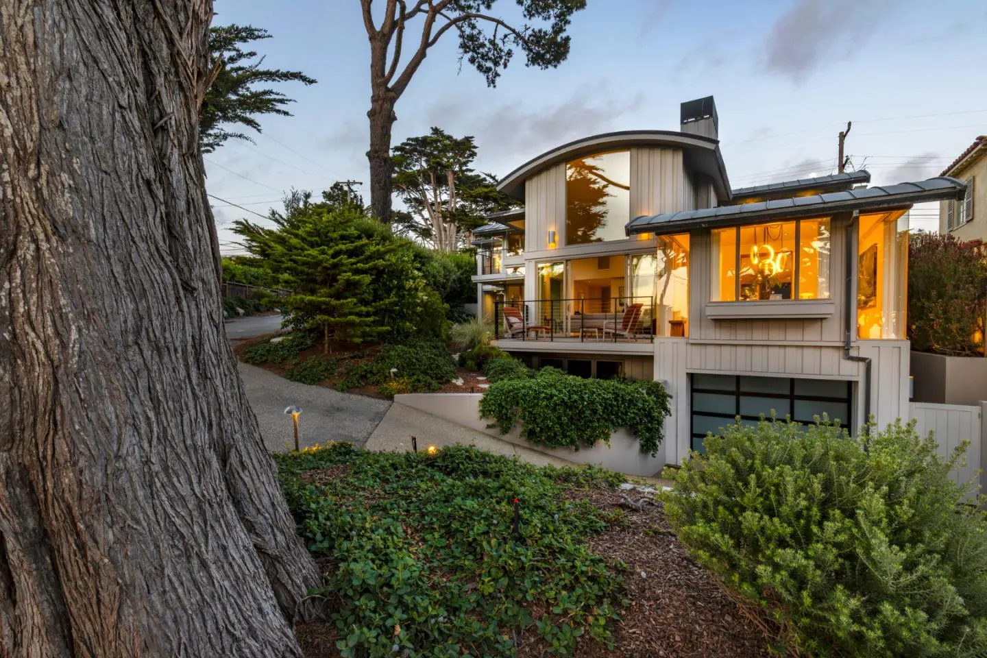 Modern two-story home with large windows and a balcony, surrounded by lush greenery and a large tree at dusk.