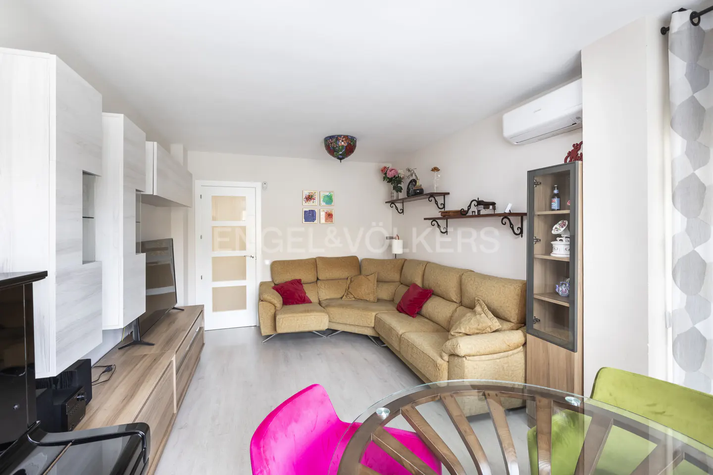 Living room with a tan sectional sofa, red pillows, glass table, pink and green chairs, and white walls.