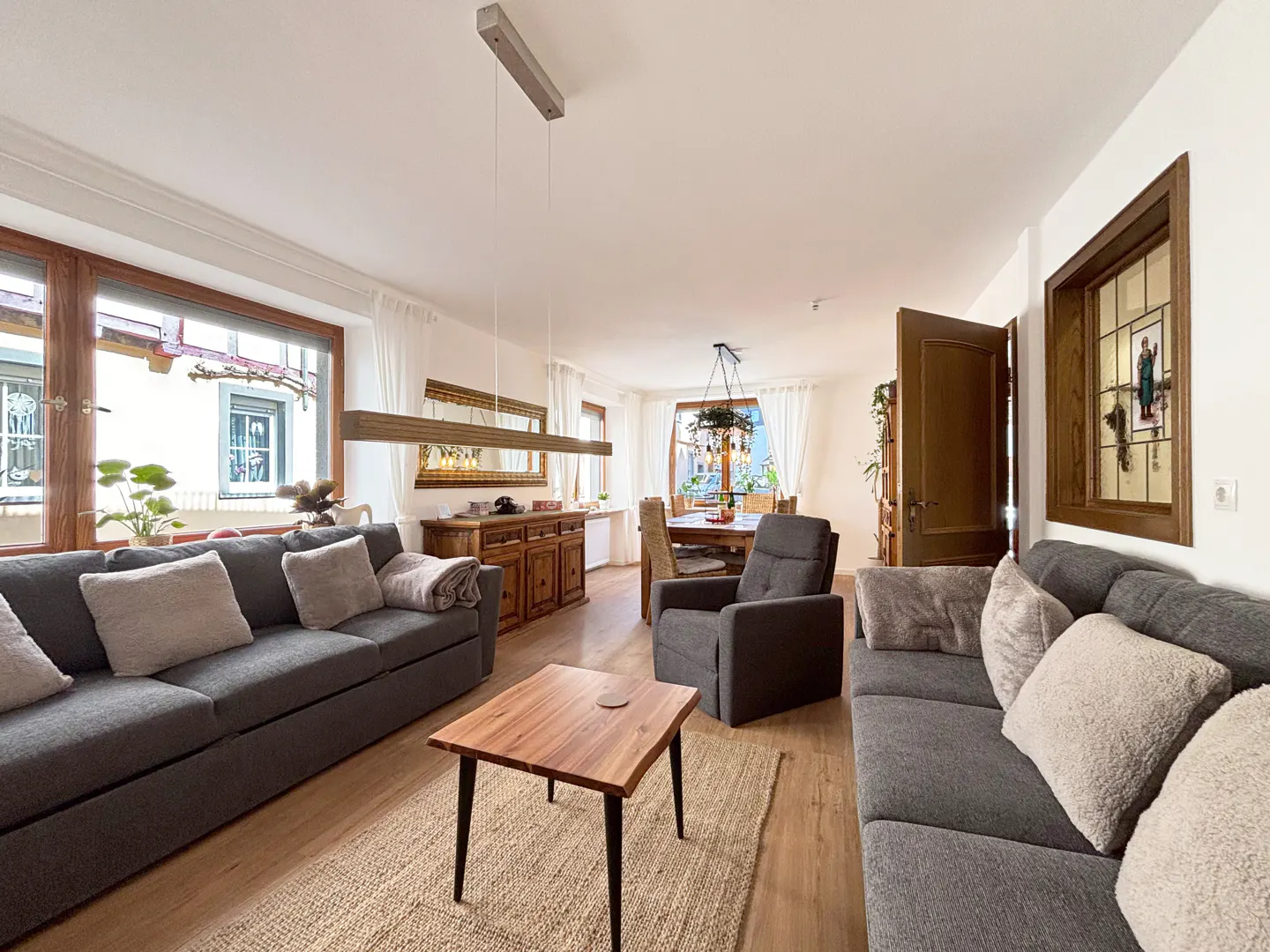 Living room with gray sofas, wood floors, and a dining area in the background. A wooden coffee table sits on a jute rug. Natural light fills the space.
