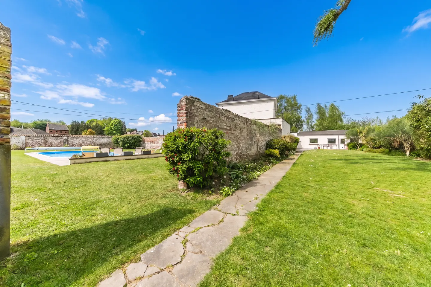 A backyard with a pool, stone wall, and a path leading to a white building under a blue sky.