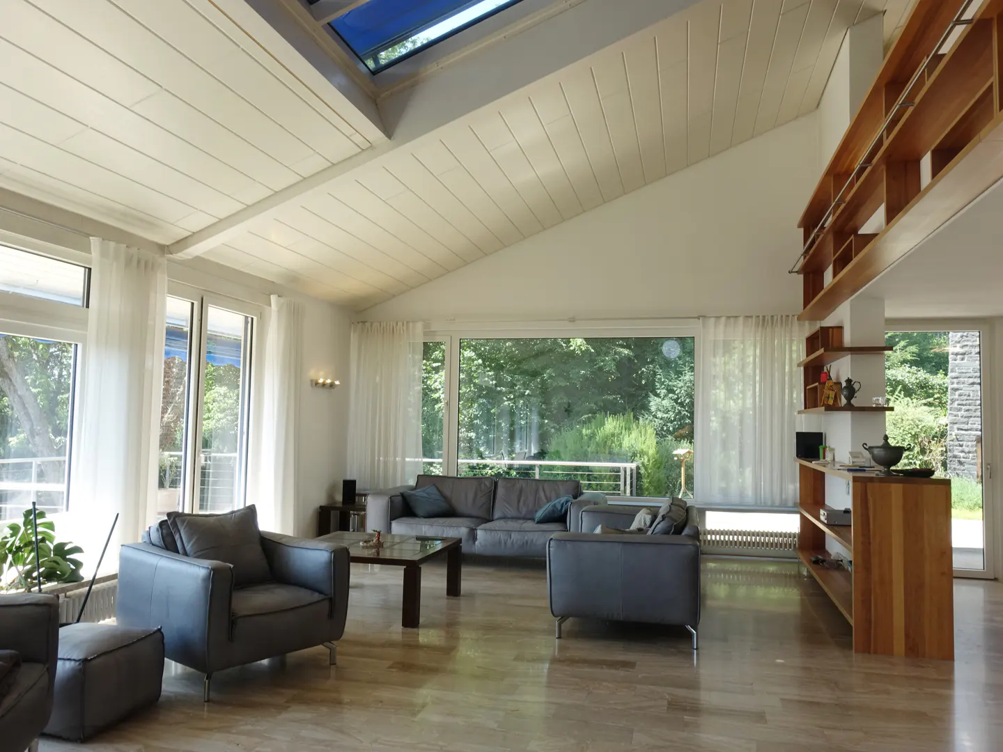 Bright living room with vaulted ceiling, skylight, grey leather furniture, and large windows overlooking green trees.