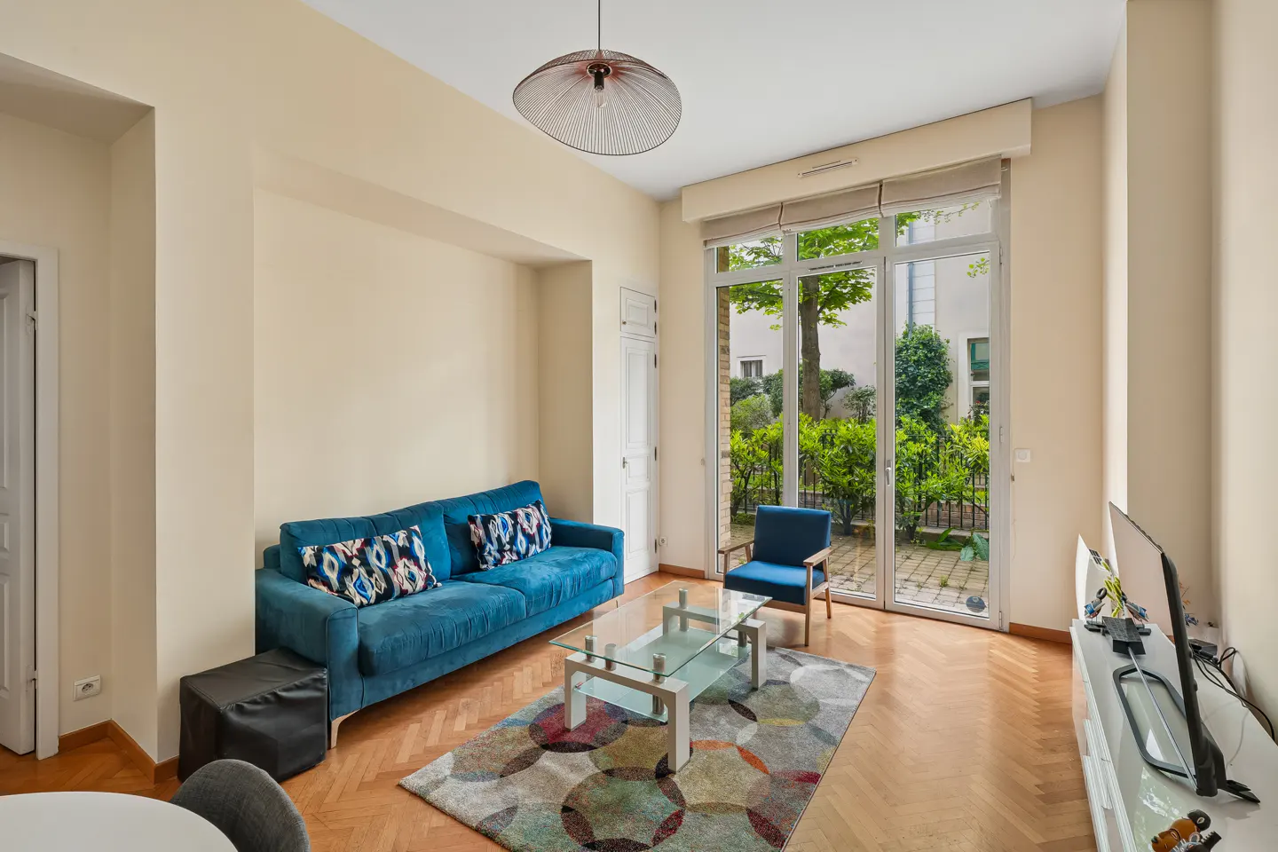 Living room with blue sofa, glass table, and view of a garden through glass doors.