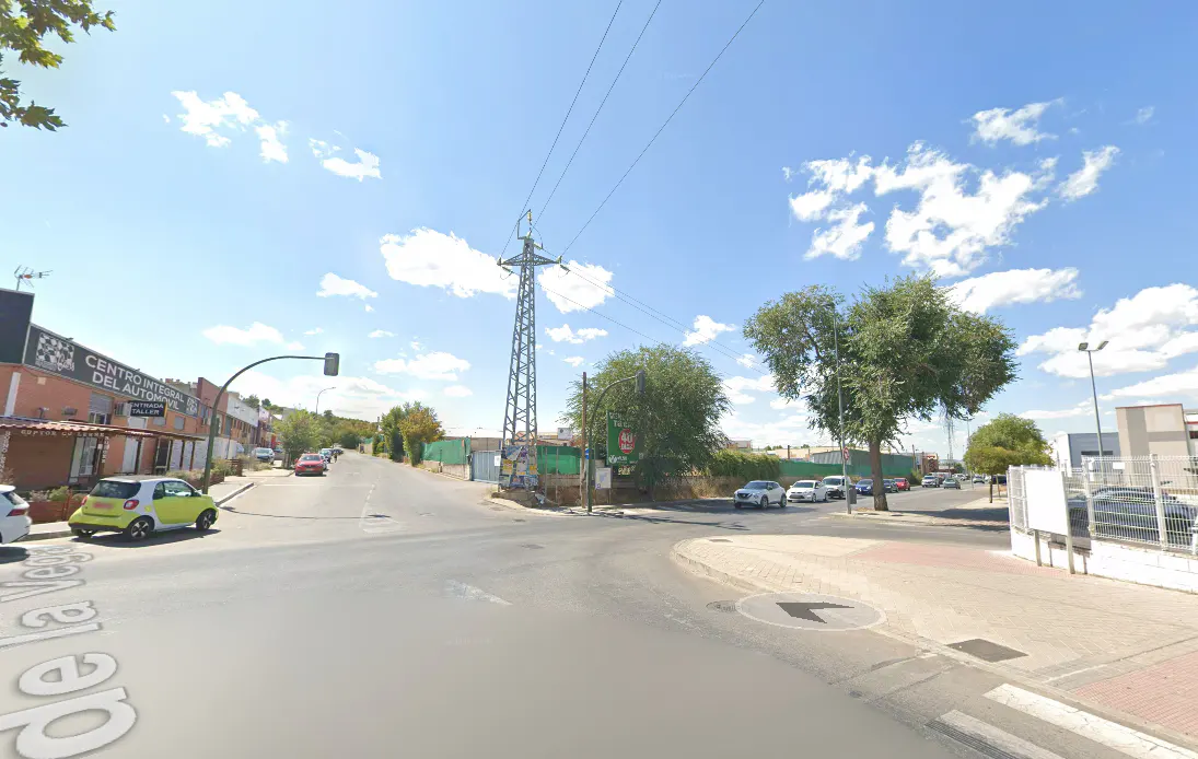 Street view of a road intersection under a blue sky with scattered clouds. Cars, buildings, trees, and a power line tower are visible.