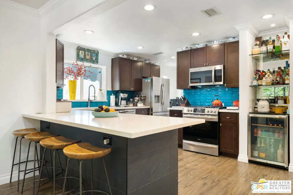 A bright kitchen with dark wood cabinets, stainless steel appliances, and a blue tile backsplash. A quartz countertop island has four wooden stools.