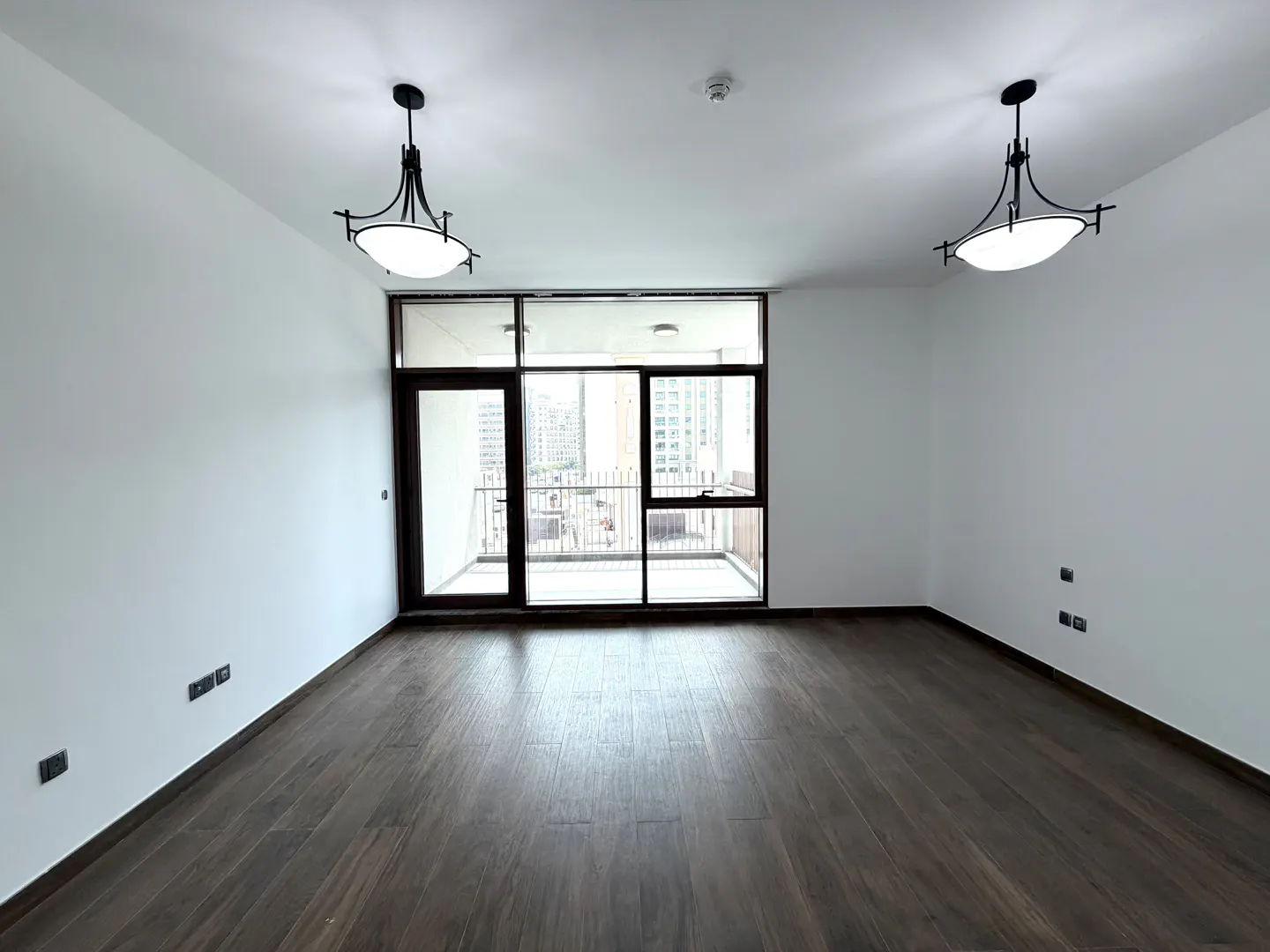 Empty room with dark wood floors, white walls, and a balcony with a city view. Two black light fixtures hang from the ceiling.