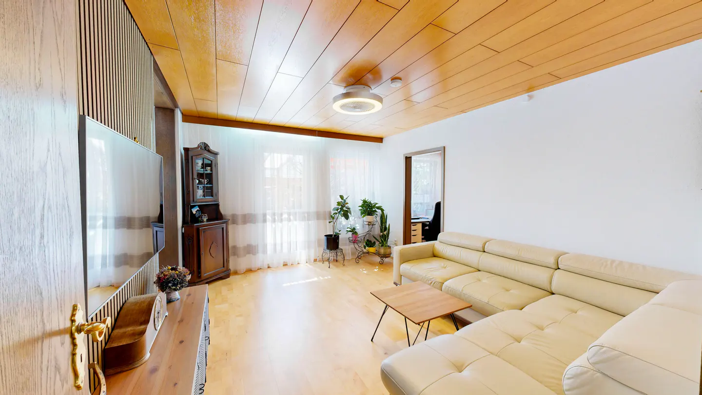 Bright living room with wood-paneled ceiling, white walls, and light wood floors. A cream-colored sectional sofa faces a TV. Plants sit near the window.