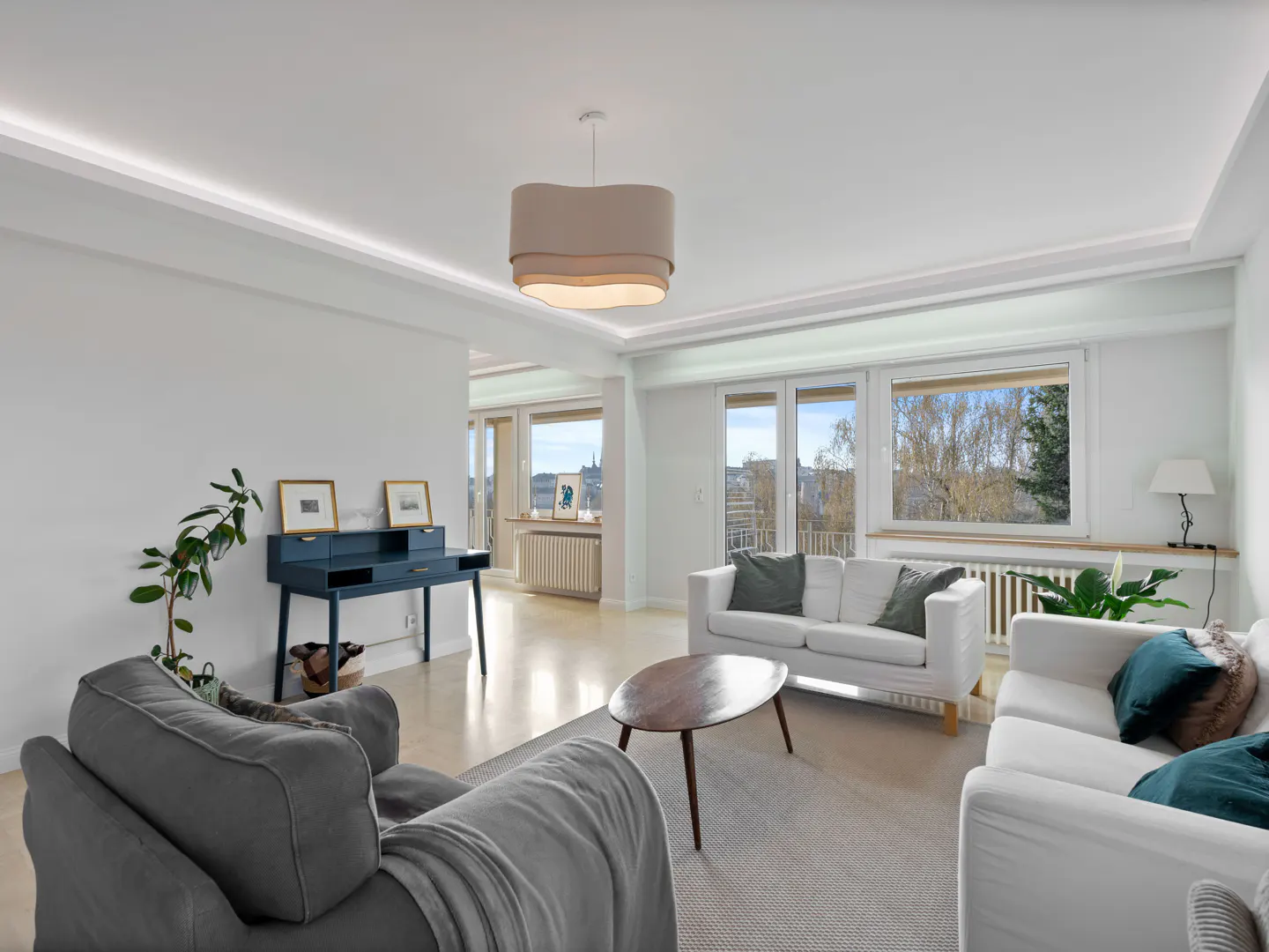 Bright living room with white walls, gray and white sofas, a wooden coffee table, and a blue desk by a large window.
