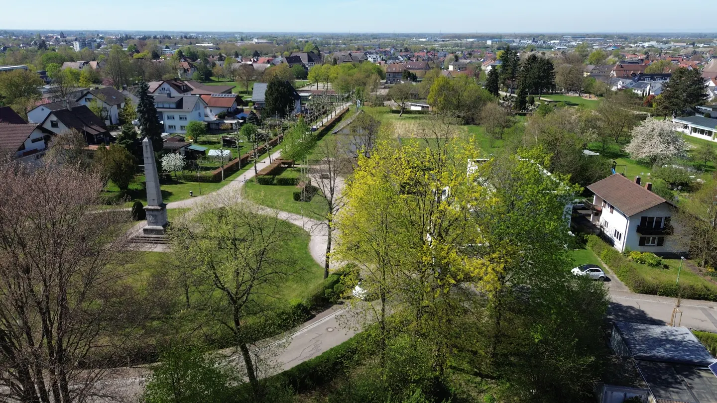 Aerial view of a park with green trees, walking paths, a monument, and houses in the background.