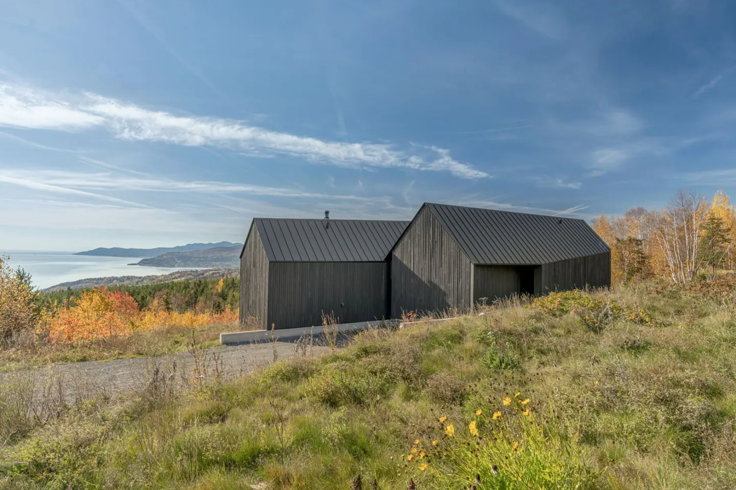 Modern black house with a metal roof on a grassy hill overlooking a bay and fall foliage.