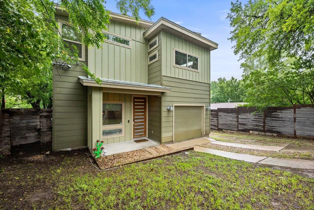 Two-story house with green siding, a modern front door, and a garage. The lawn is green and there are trees in the background.