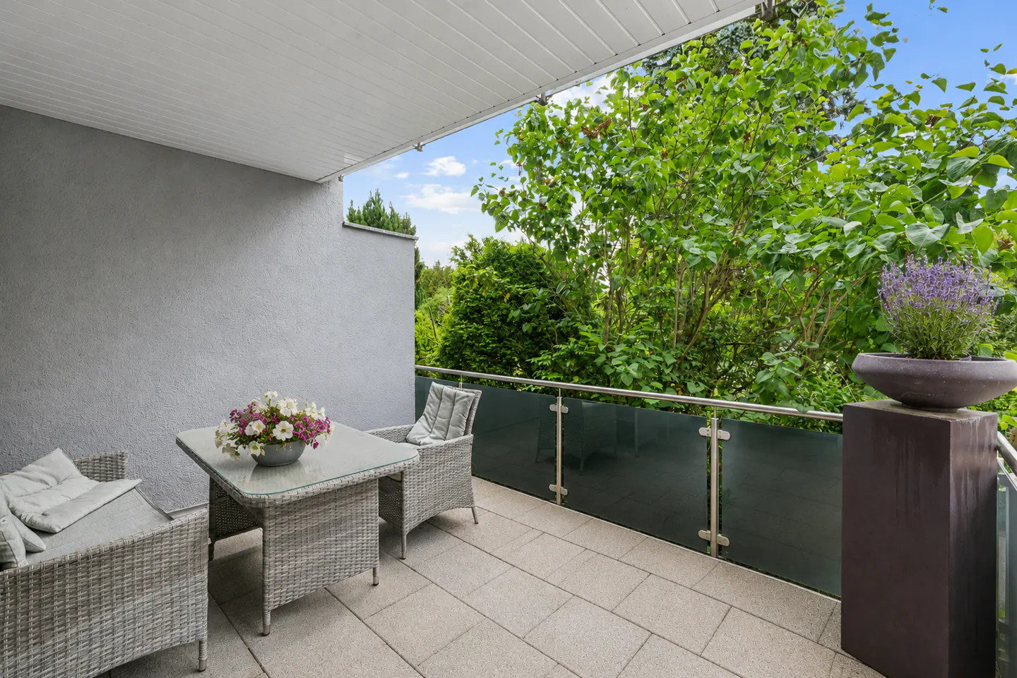 A covered balcony with gray wicker furniture, including a table with flowers, chairs, and a view of lush green trees.