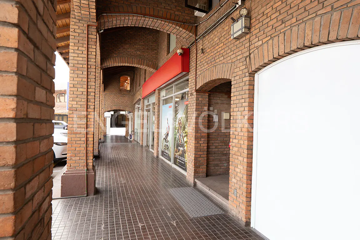 Brick arcade with storefronts. Red awnings above glass windows. Gray tiled floor. Arched brick openings lead to more shops.