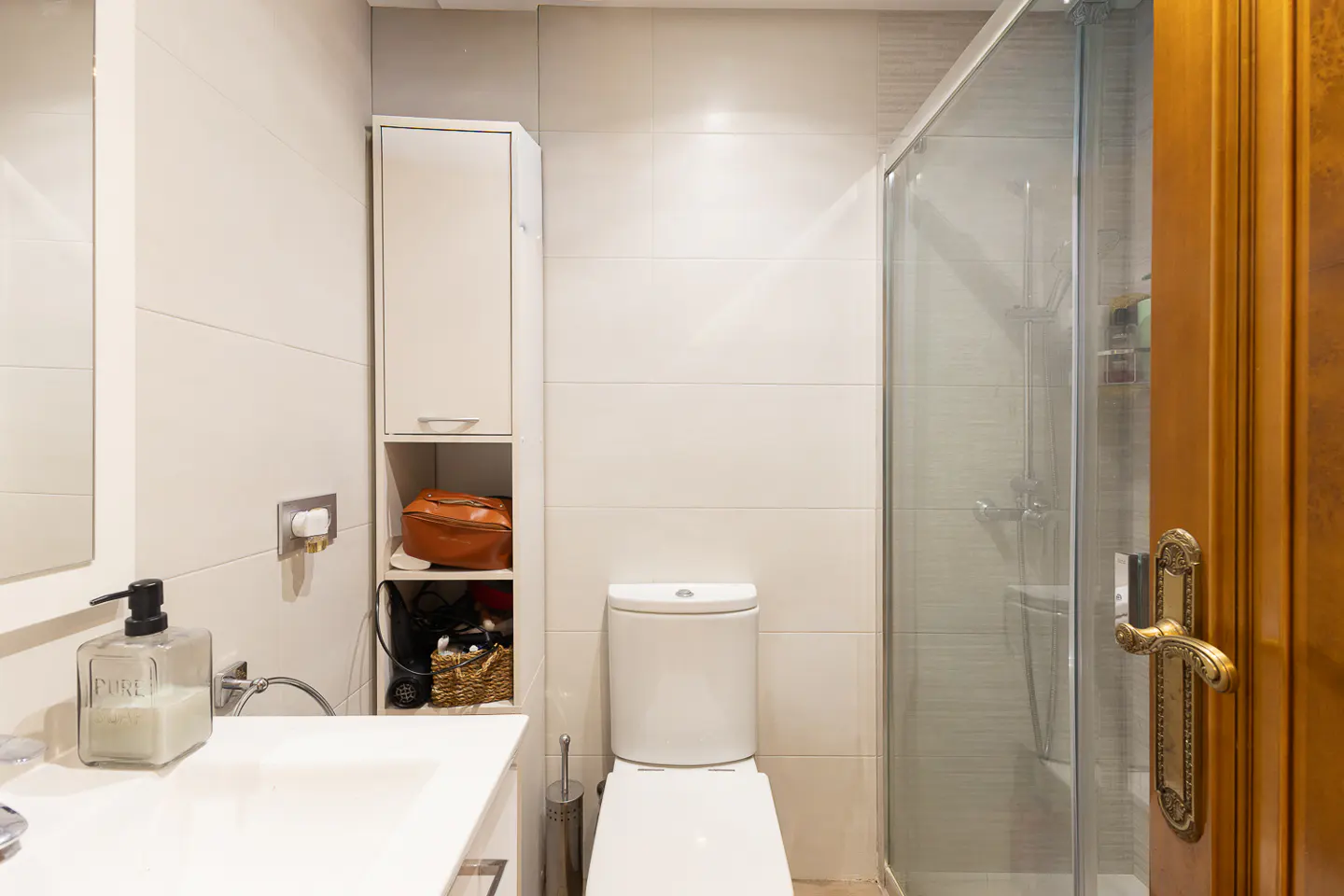 Modern bathroom with white tiles, a toilet, a glass shower stall, a vanity with soap, and a tall storage cabinet.