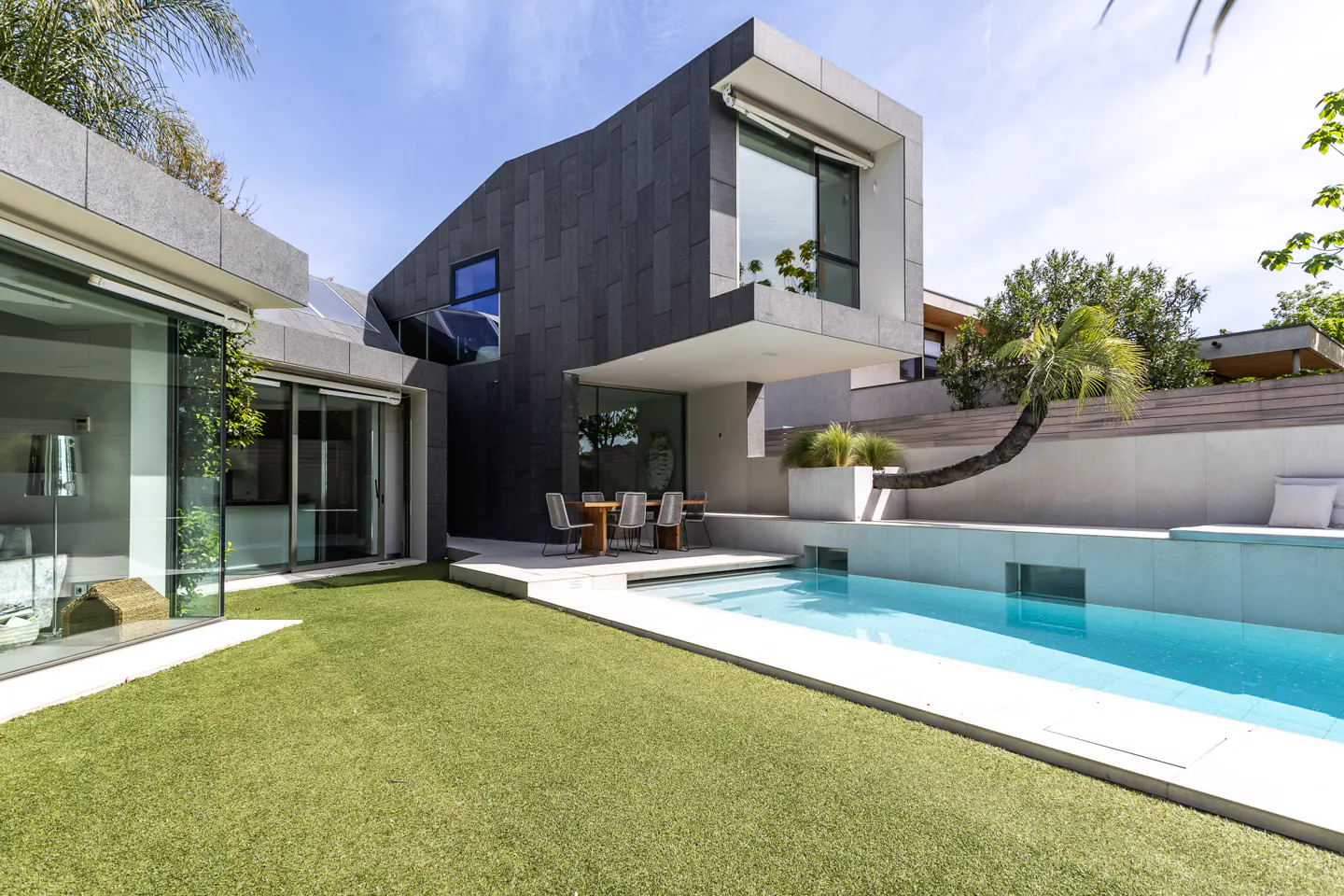 Modern home exterior with a pool, green lawn, and outdoor dining. The house is gray and white with large windows.