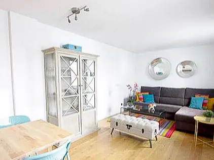 Bright living room with white walls, wood floors, and a gray sectional sofa with colorful pillows. A white cabinet stands near a light wood dining table.