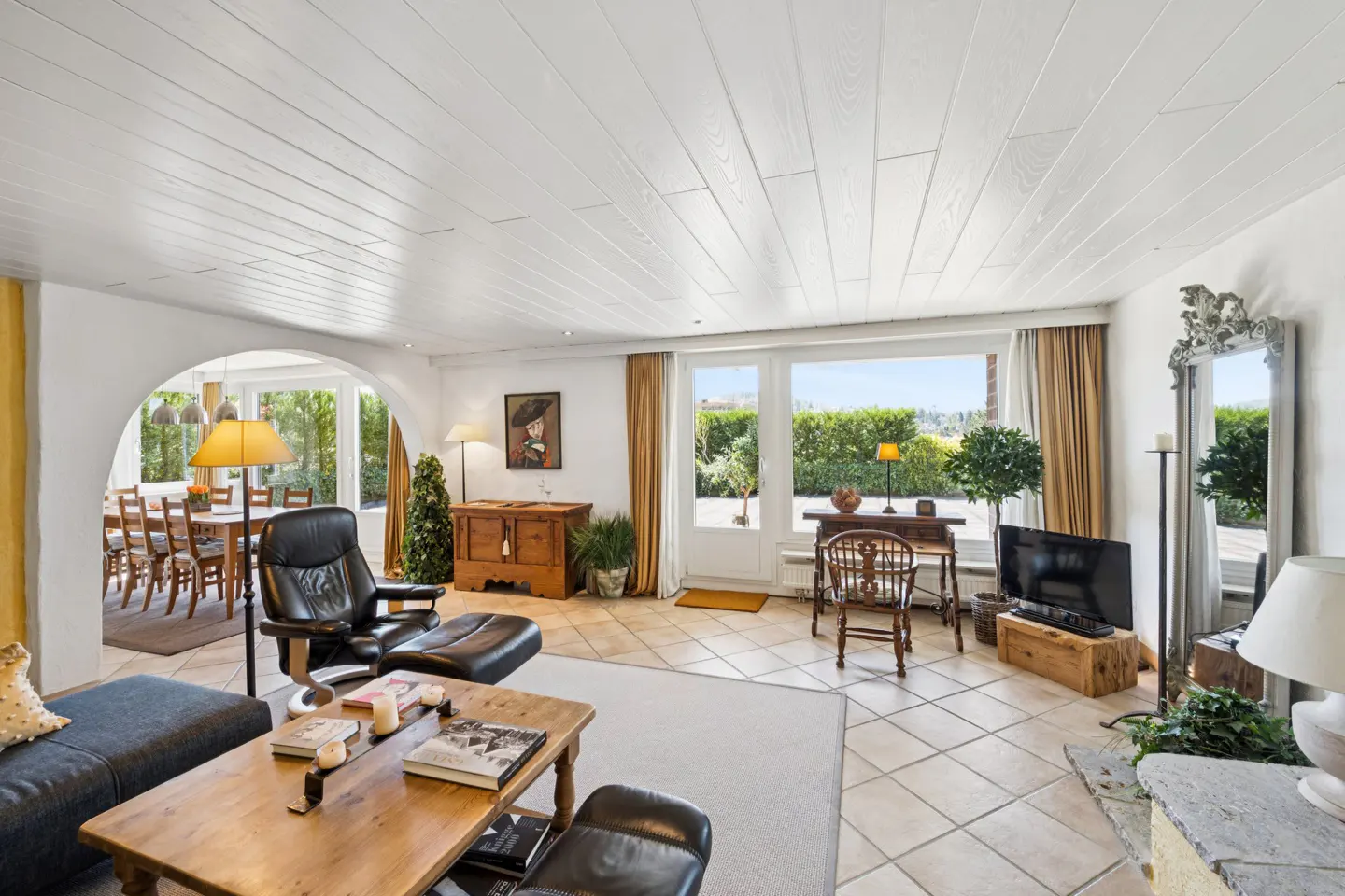 Bright living room with white wood ceiling, tile floor, leather chairs, wood furniture, and large windows overlooking green hedges.