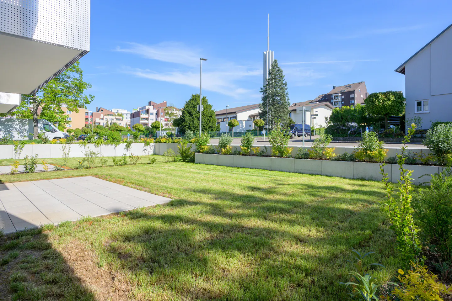 A green lawn with a concrete patio, bordered by a low concrete wall with shrubs, in a residential area under a blue sky.