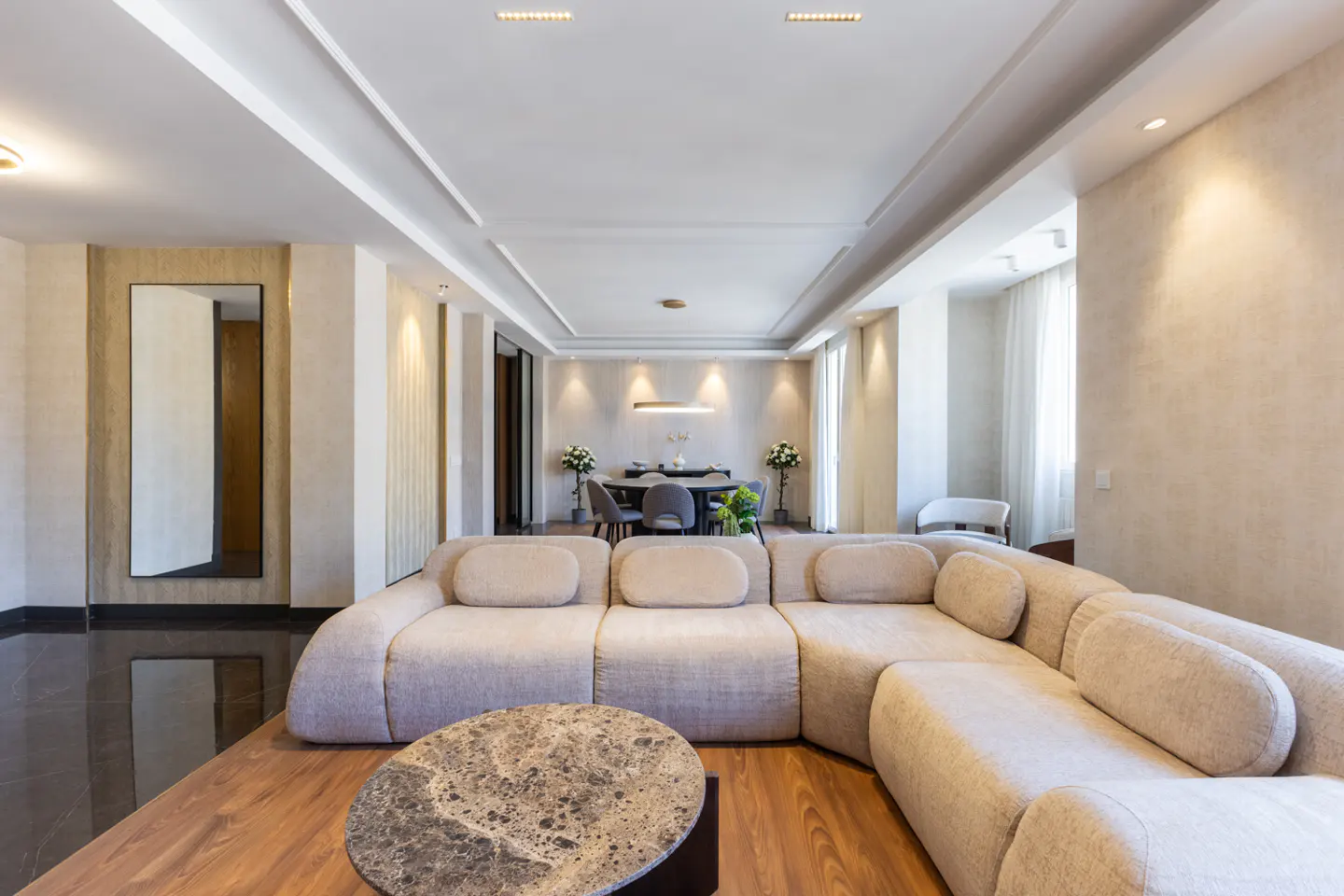 Modern living room with a large beige sectional sofa, a round marble coffee table, and a dining area in the background.