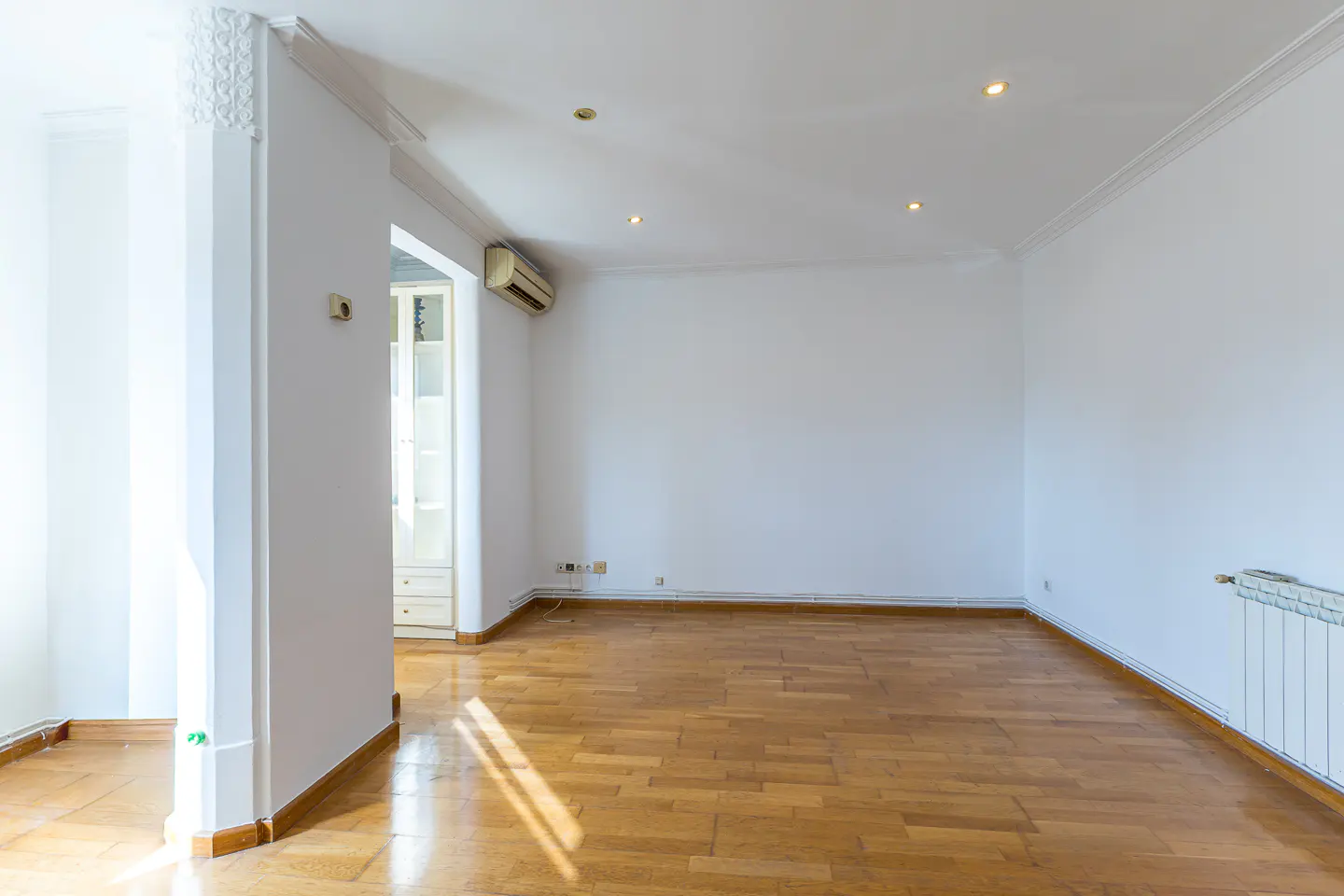 Bright, empty room with hardwood floors, white walls, and ceiling. A doorway leads to a room with a white cabinet. An air conditioner is mounted on the wall.