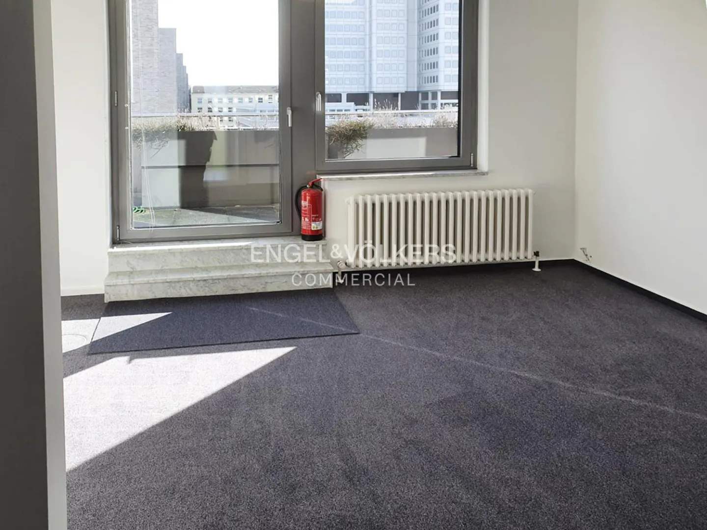 Bright, empty office space with gray carpet, white walls, and a large window overlooking city buildings. A red fire extinguisher is visible.