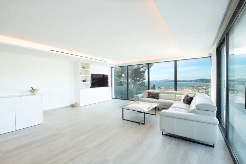 Bright, modern living room with white walls, tile floor, and large windows overlooking the ocean. A white sectional sofa and marble coffee table are featured.