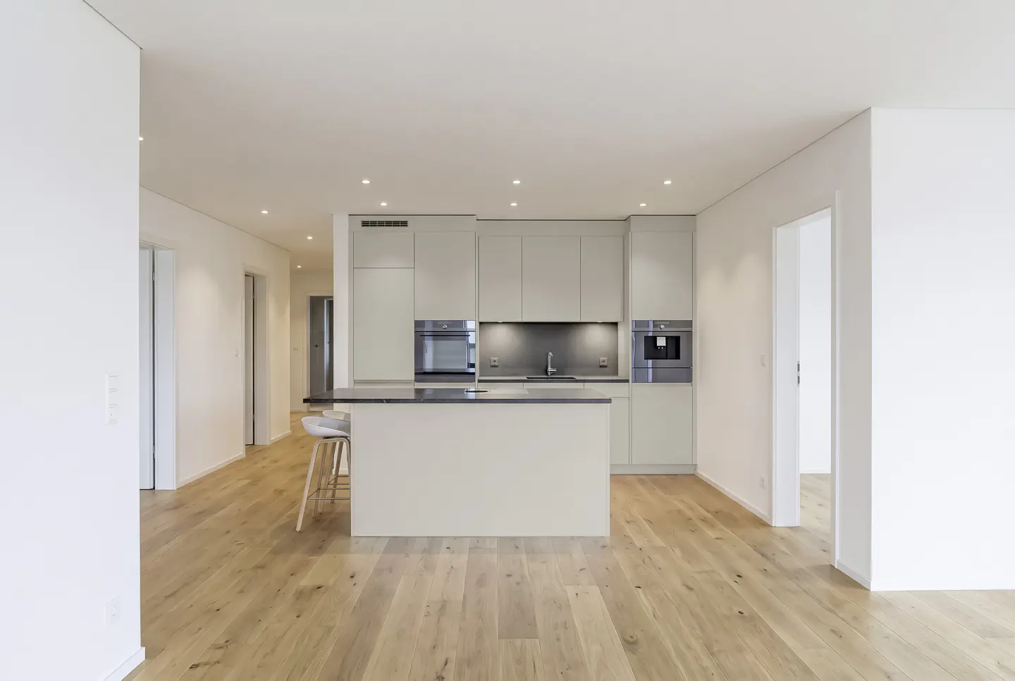 Modern kitchen with light grey cabinets, a central island with bar stools, and light wood flooring in an open-plan space.